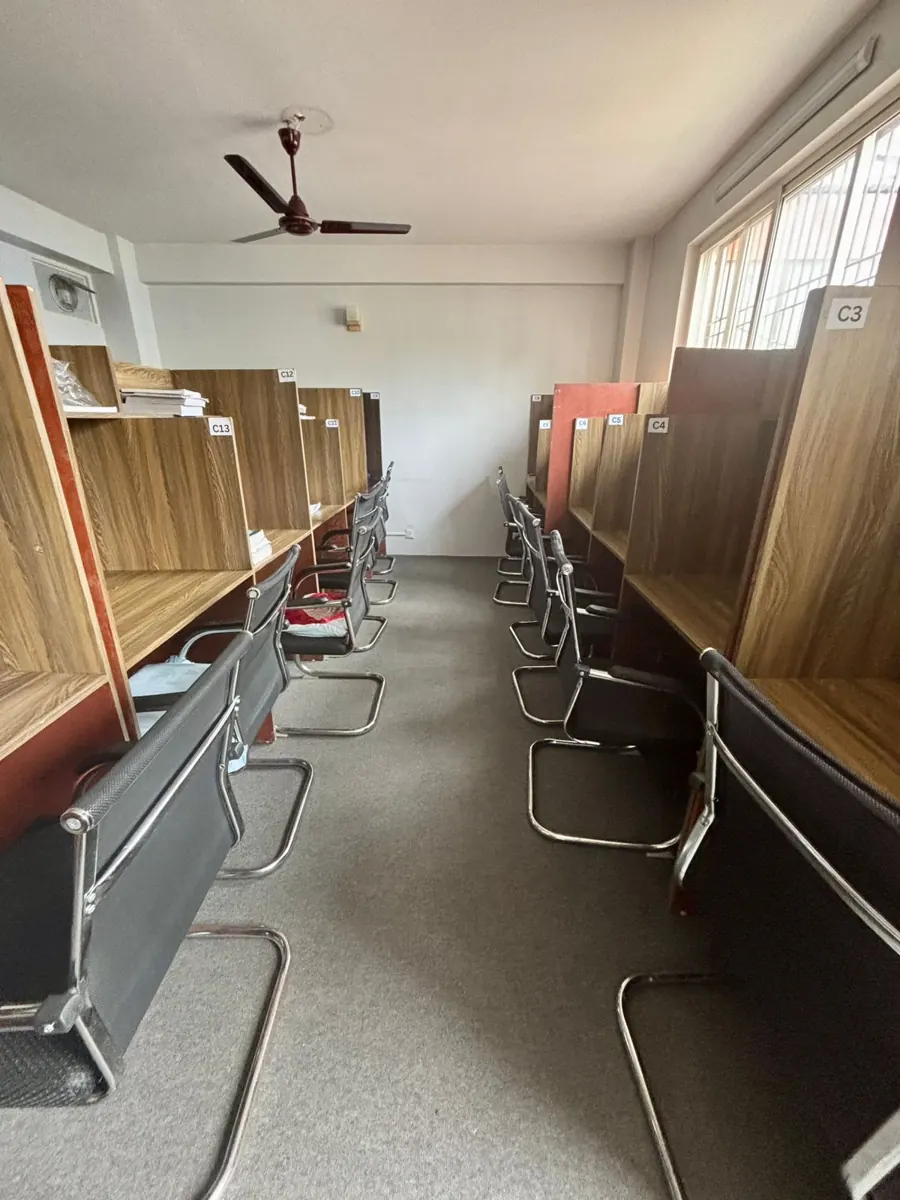 A bright hall with two rows of wooden cabins and mesh chairs under a ceiling fan.