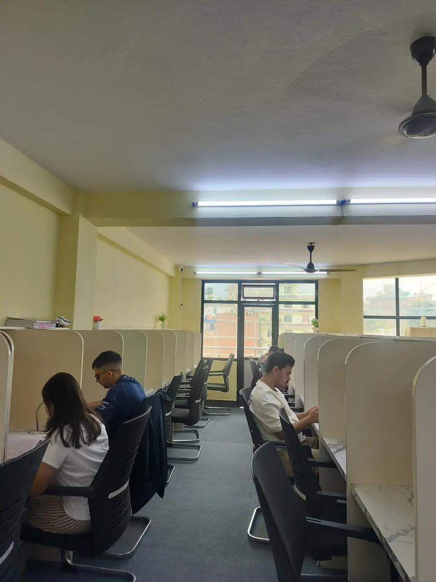 Students studying in rows of cream cabins under ceiling fans, with windows at the far end of the hall.